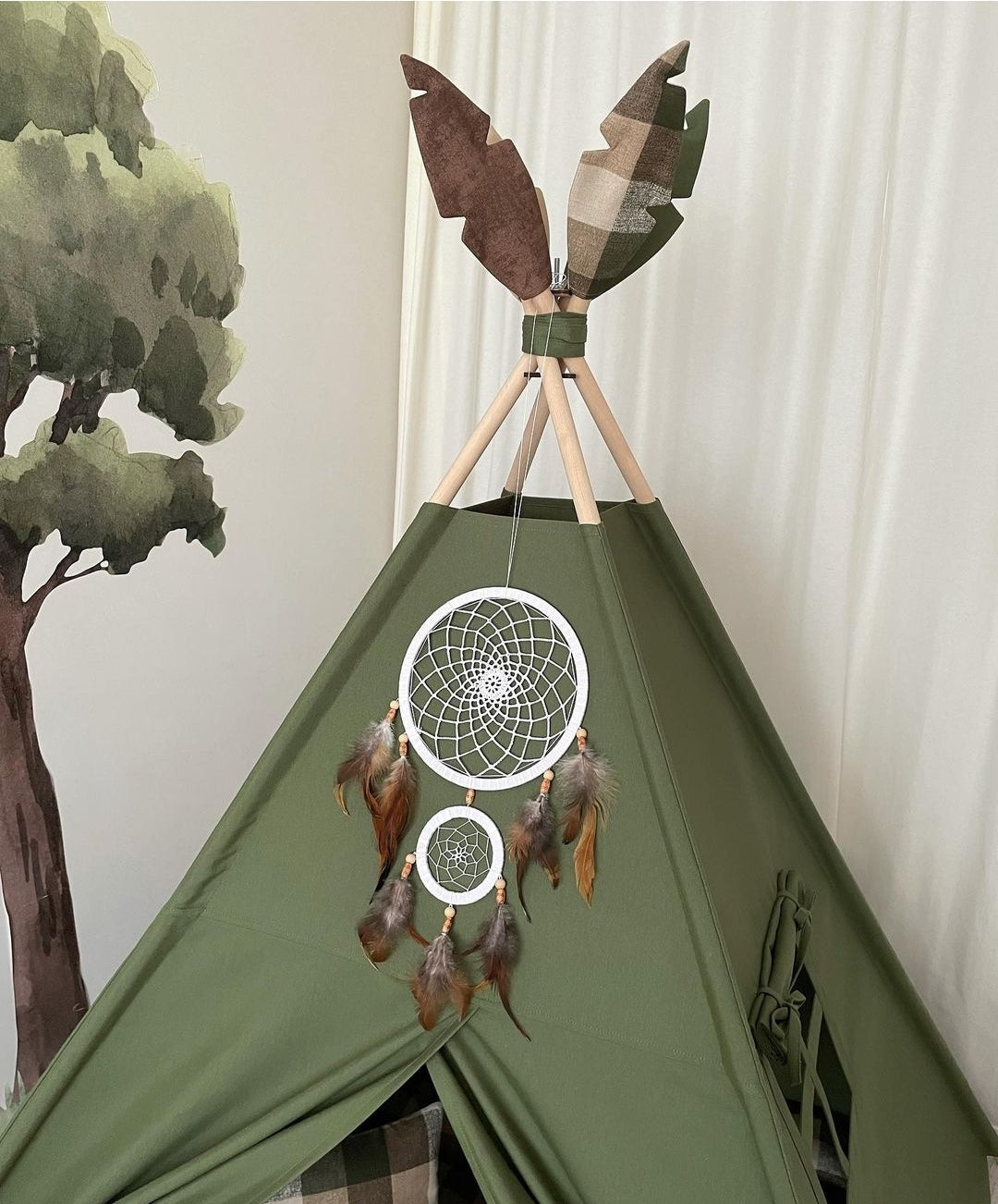 Na slici je vrh zelenog šatora sa hvatačem snova i ukrasnim perima od kojih je jedan kariran drugi je braon a treći zeleni na vrhu. Pored njega je naslikano drvo na zidu.
