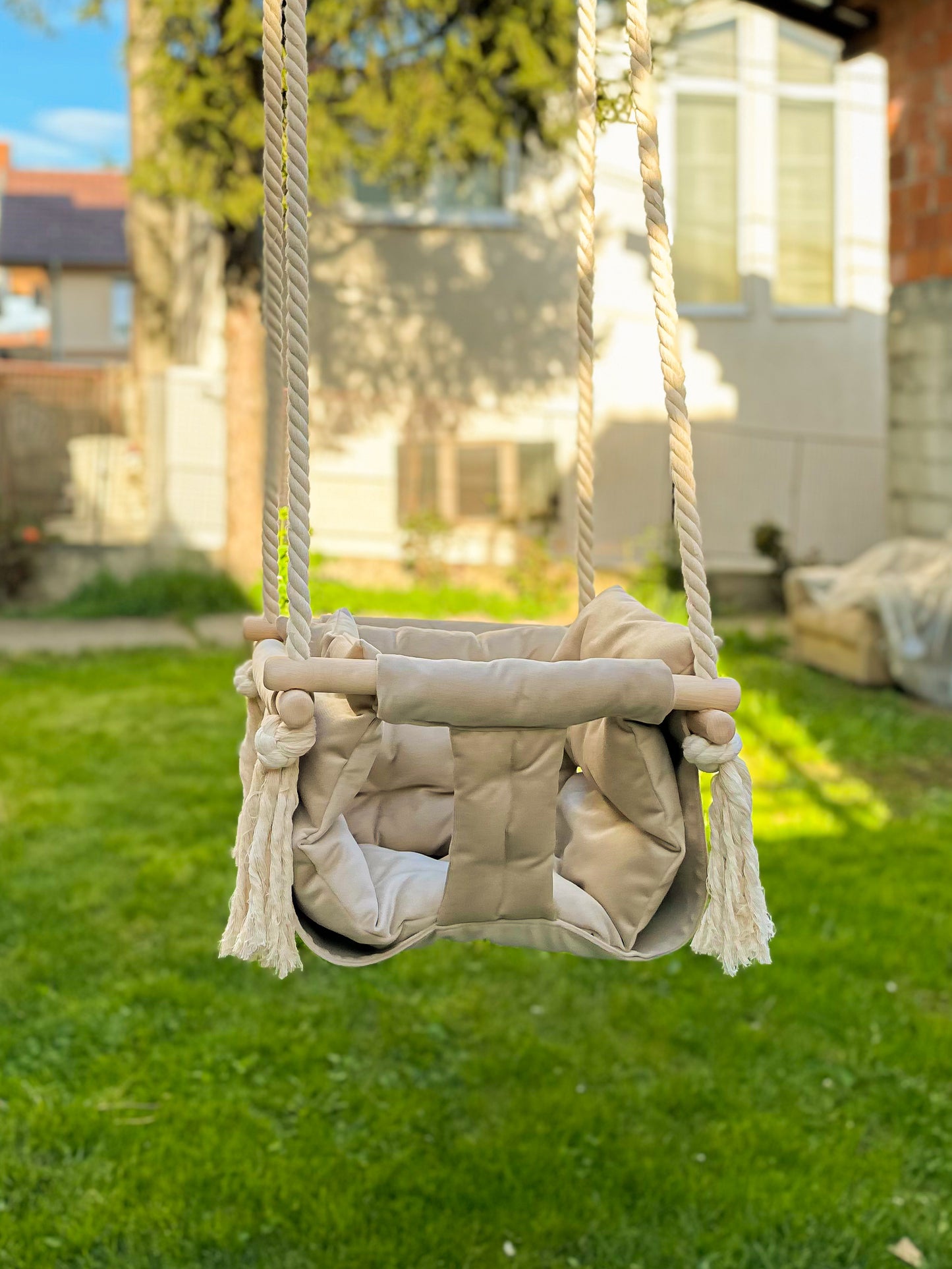 Elegantna dečija ljuljaška postavljena u predivnoj bašti, sa zelenom travom i toplom kućom u pozadini. Ljuljaška je izrađena od meke, postavljene tkanine svetle bež boje, sa dodatnim jastukom za udobnost i čvrstim drvenim okvirom. Stabilni pamučni užadi završavaju se dekorativnim čvorovima, dajući prirodan i moderan izgled. Postavljena na otvorenom, ova ljuljaška odiše toplinom i poziva na igru i opuštanje na svežem vazduhu.