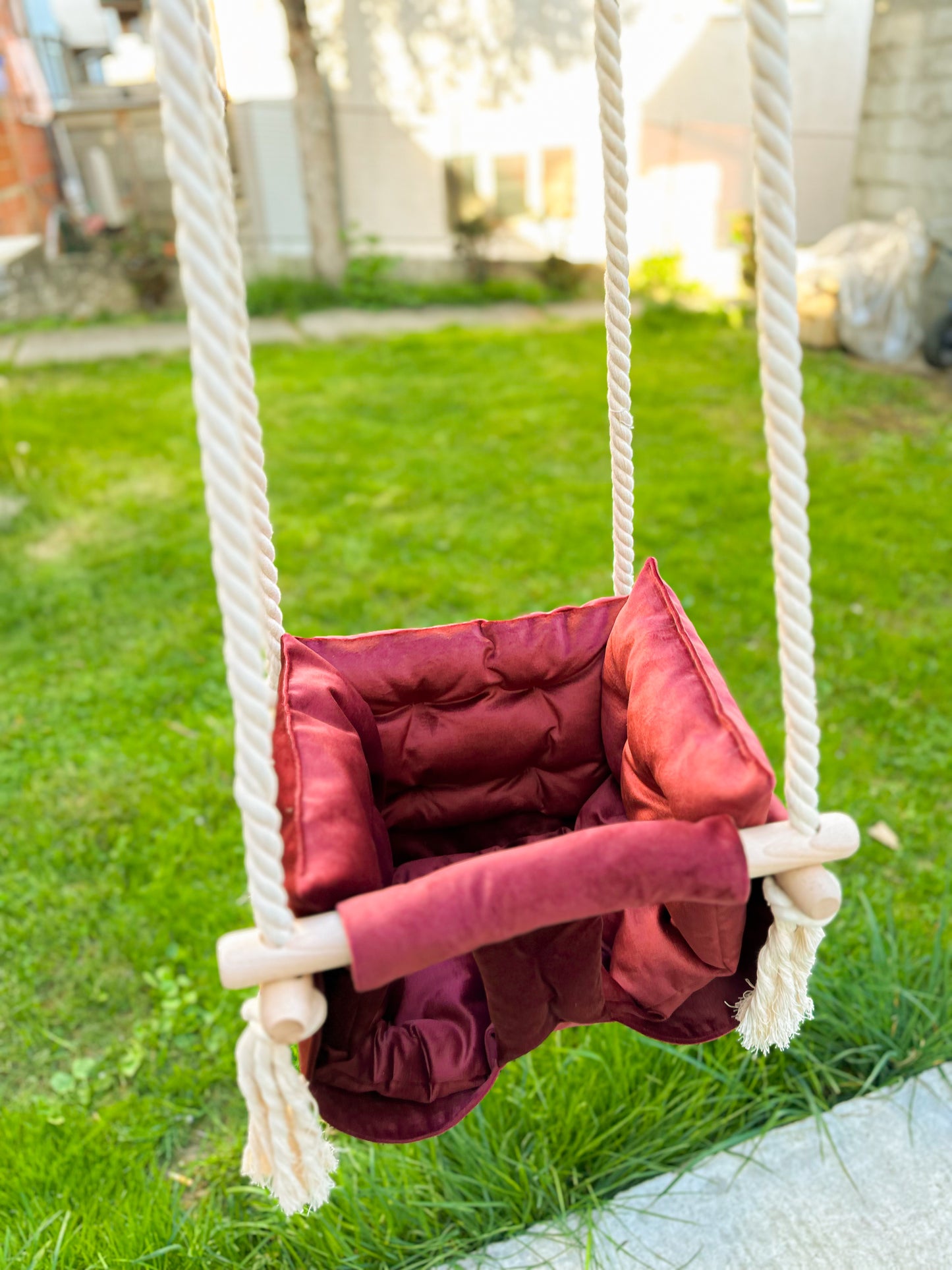 Elegantna dečija ljuljaška postavljena u predivnoj bašti, sa zelenom travom i toplom kućom u pozadini. Ljuljaška je izrađena od meke, postavljene tkanine bordo boje, sa dodatnim jastukom za udobnost i čvrstim okvirom od drveta. Debelih pamučni užadi završavaju se dekorativnim čvorovima, dajući moderan izgled. Postavljena na otvorenom, ova ljuljaška odiše toplinom i poziva na igru,radost i opuštanje na svežem vazduhu.