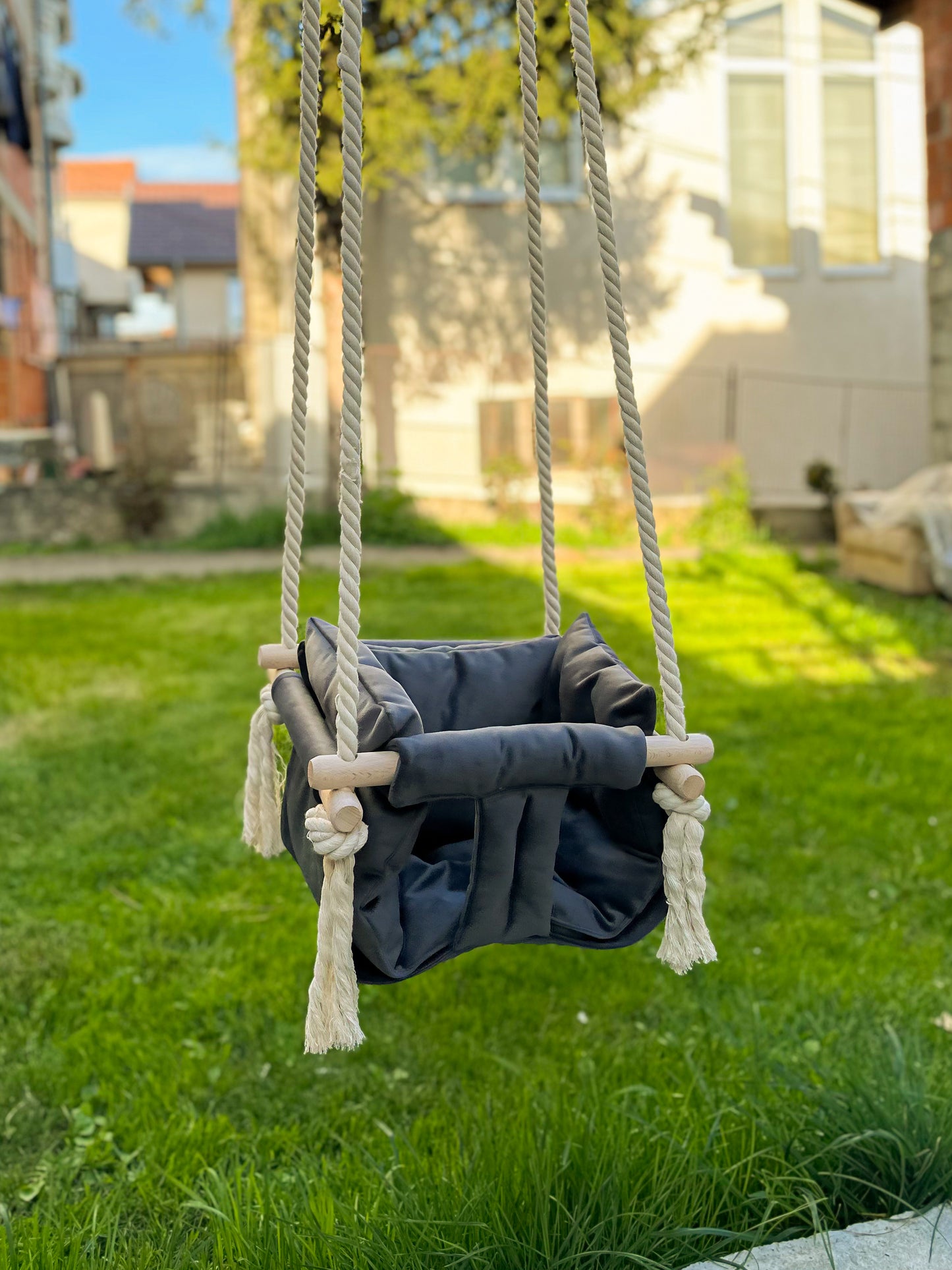 Ljuljaška za baštu u sivoj nijansi, postavljena u avliji sa bujnom travom u pozadini. Napravljena je od nežnog tekstila sa drvenim držačima, a visi na čvrstim svetlim užadima ukrašenim resicama. Fotografija je snimljena u prirodnom okruženju tokom sunčanog dana, što naglašava prijatan kontrast između ljulajške i osvetljenog travnjaka. Savršena je za igru na otvorenom, kombinujući funkcionalnost i atraktivan dizajn. Svojim stilom se skladno uklapa u baštenski ambijent, pružajući deci razlog za igru.