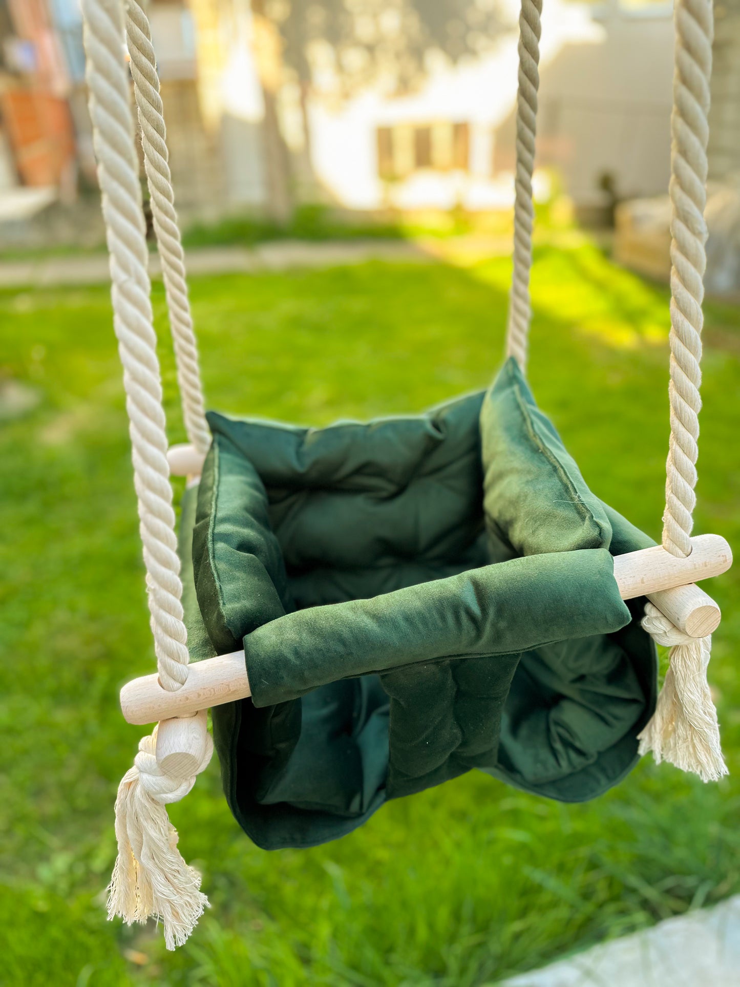 Ljuljaška za baštu u zelenoj boji, postavljena u avliji sa travom u pozadini. Napravljena je od tekstila sa drvenim držačima, a visi na čvrstim svetlim užadima ukrašenim resicama. Fotografija je snimljena u prirodnom okruženju tokom sunčanog dana, što naglašava prijatan kontrast između ljulajške i osvetljenog travnjaka. Savršena je za igru na otvorenom. Svojim stilom se skladno uklapa u baštenski ambijent, pružajući deci razlog za igru.