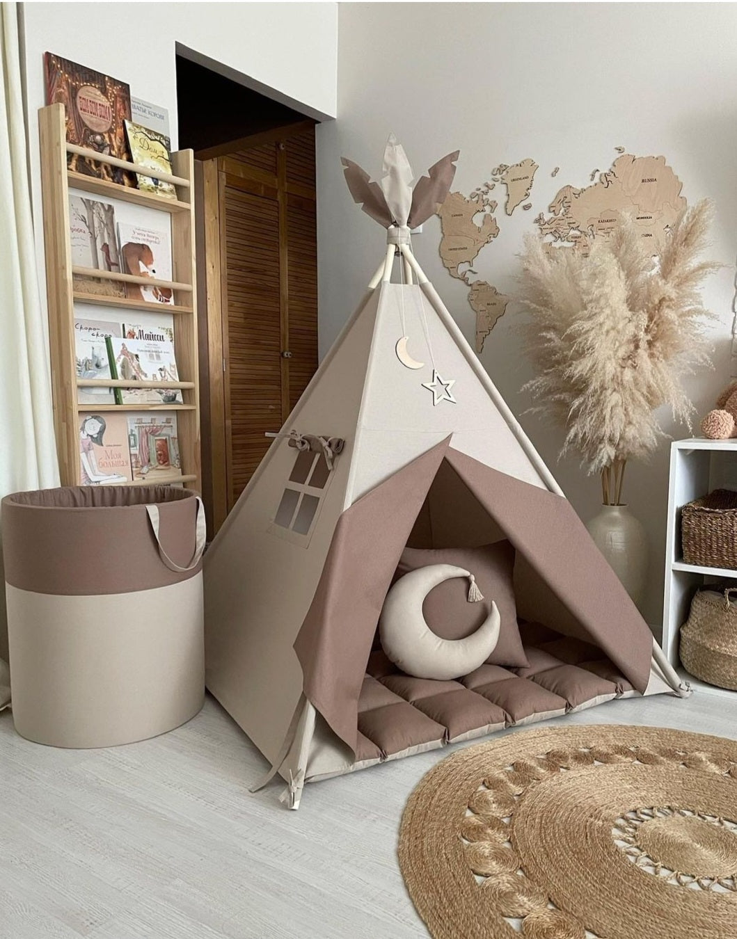 Estetika slike gde dominiraju prirodni materijali i smirujuće boje. Smeđi šator za decu u kombinaciji s prostirkom i jastukom u obliku polumeseca stvara udoban i privlačan kutak za igru ili odmor. Tepih unosi prirodnu toplinu u prostor, dok dekoracije poput suve trave i drvene mape sveta na zidu dodaju dozu sofisticiranosti. Police s knjigama u drvenom okviru pružaju funkcionalnost, dok svetle boje zidova i poda naglašavaju prostranost i jednostavnost prostora.
Pored šatora je kanta koja je bež i braon boje
